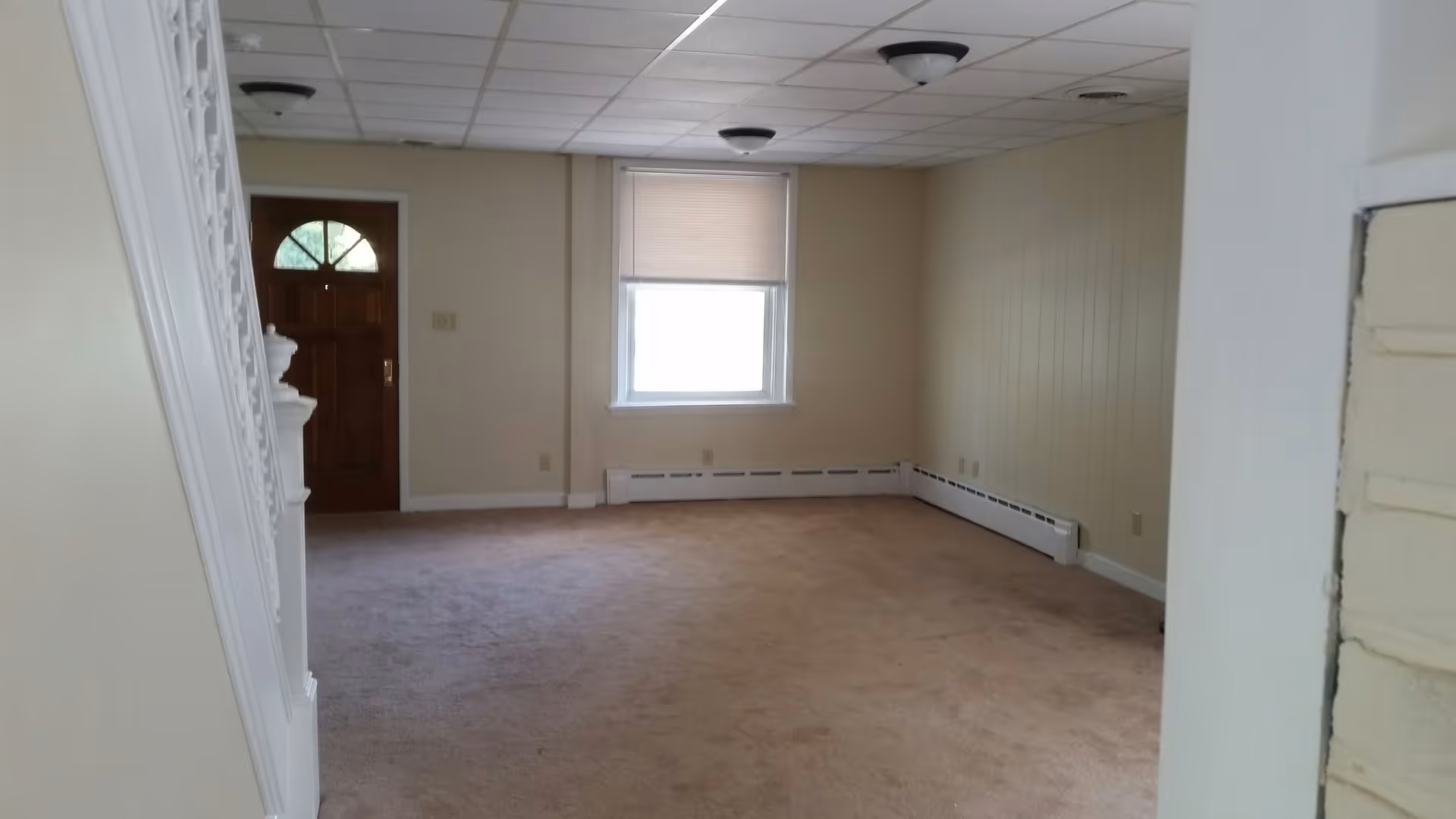 Empty living room with beige carpet and walls, a window with a closed blind, a wooden front door with a glass panel, and a white staircase railing on the left side.