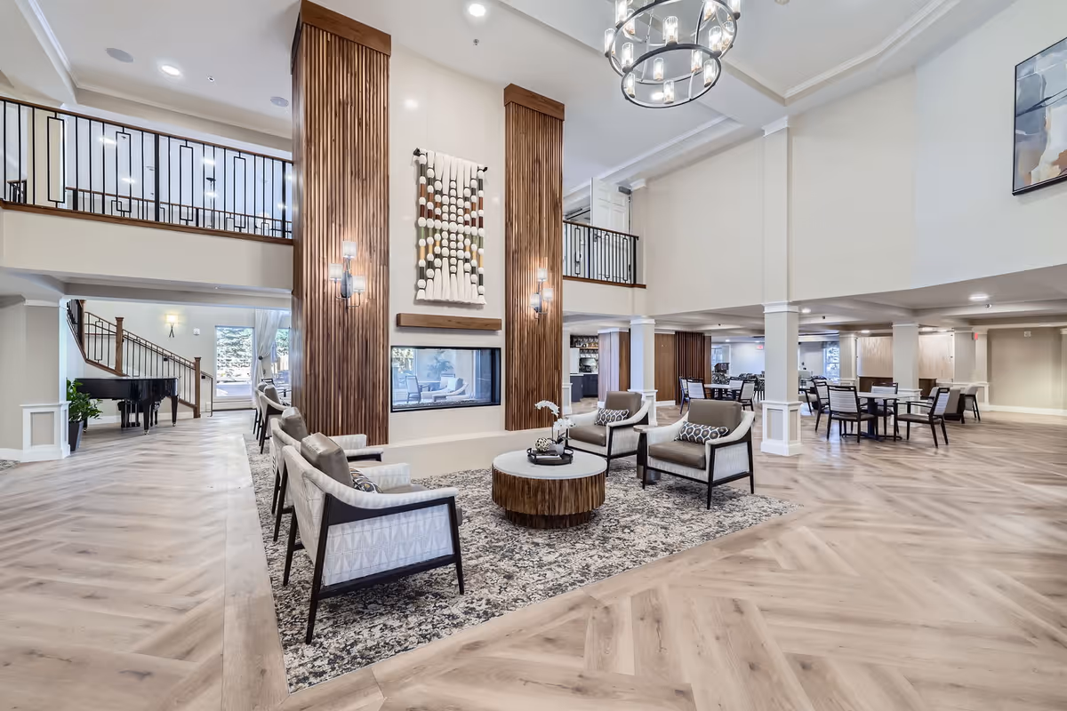 Spacious and modern common area in a retirement community featuring a seating arrangement with four armchairs around a round coffee table on a patterned rug. The room has a high ceiling with a large chandelier, wooden columns with wall sconces, and a decorative wall hanging above a built-in fireplace. In the background, there are dining tables and chairs, a grand piano near a staircase, and large windows letting in natural light.