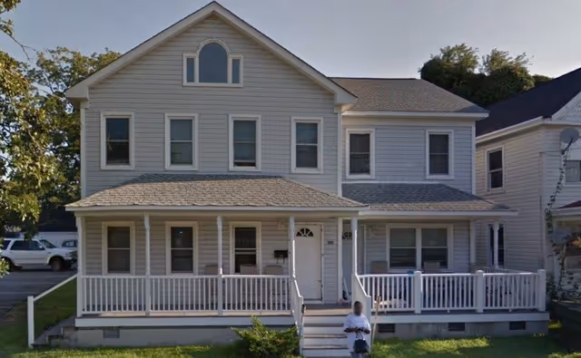 Front exterior view of a two-story light gray house with white trim and a covered porch with white railings. There are multiple windows on both floors and a person standing on the front steps. Trees and neighboring houses are visible in the background.