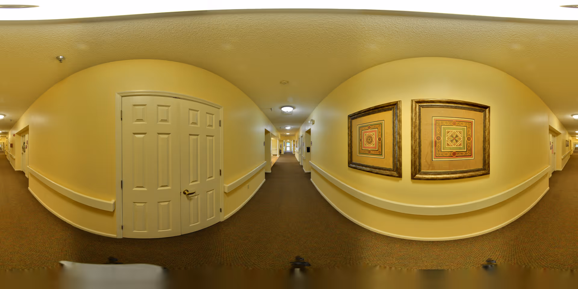 A long indoor hallway with beige walls and carpeted floor in a senior living facility. The hallway features handrails on both sides, several closed doors, and two framed decorative artworks on the right wall. Ceiling lights illuminate the corridor.