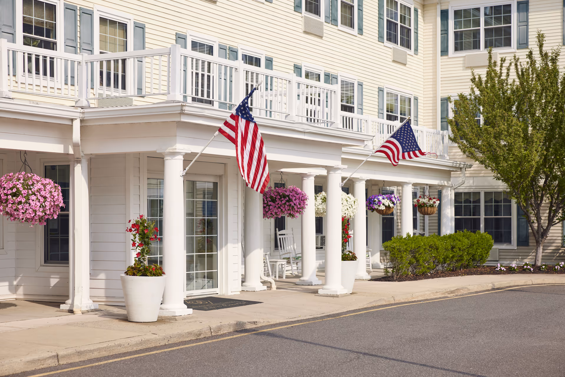 Exterior view of a senior living facility with white columns supporting a covered walkway. American flags are displayed on poles attached to the building. Hanging flower baskets and potted plants decorate the entrance area. The building has multiple windows with blue shutters and a tree and shrubs are visible on the right side.