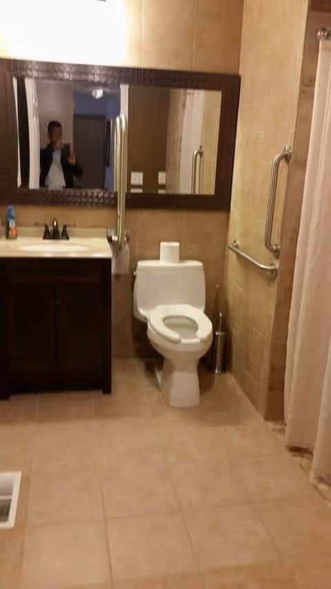 A bathroom with beige tiled walls and floor, featuring a white toilet with a toilet paper roll on top, a dark wood vanity with a sink and faucet, a large rectangular mirror above the sink, and metal grab bars on the walls near the toilet and shower area. A person is partially visible in the mirror taking the photo.