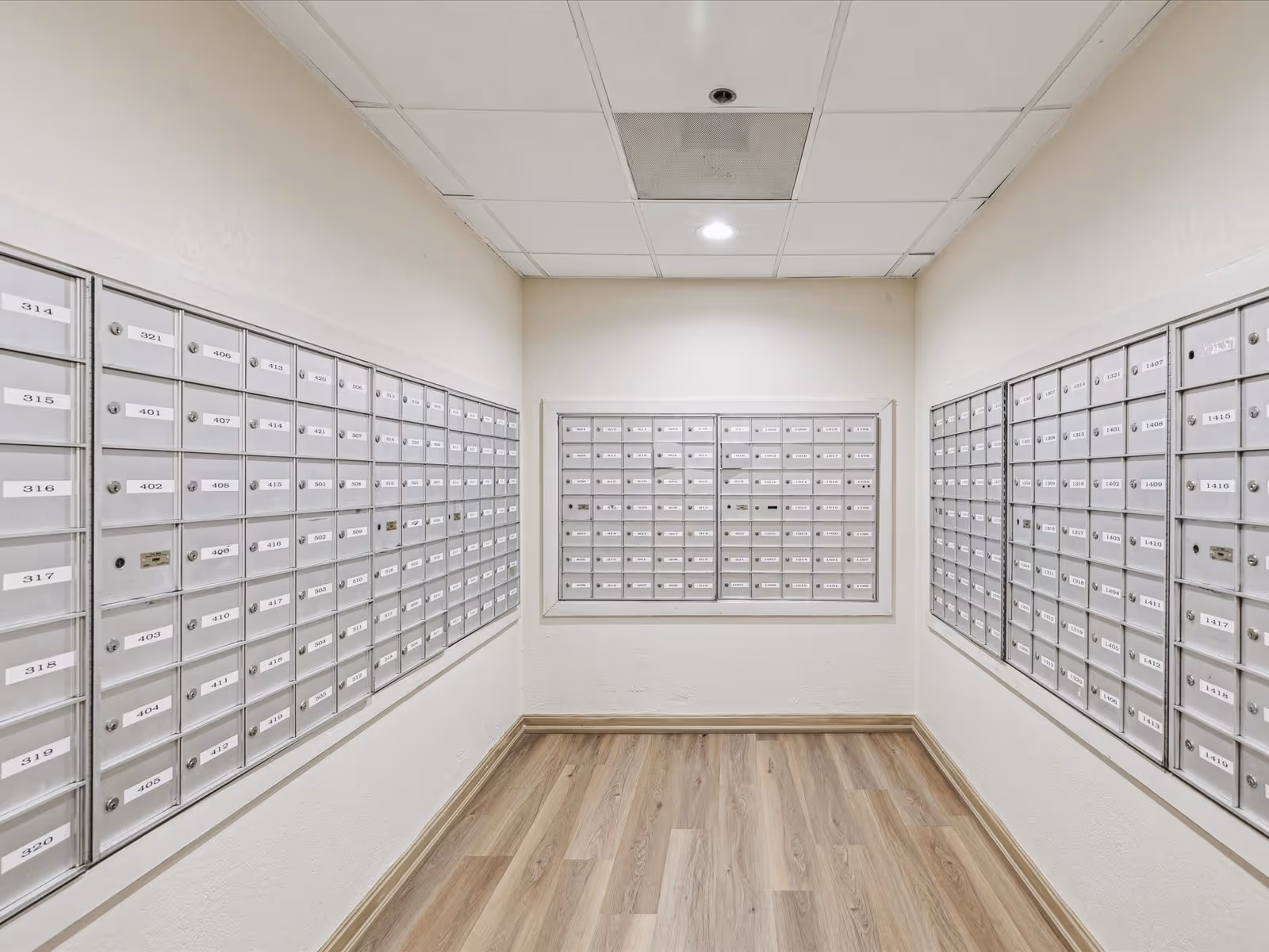 A small mailroom with three walls covered in rows of silver mailboxes, each labeled with numbers. The room has light-colored walls, a wood-patterned floor, and a white ceiling with recessed lighting.