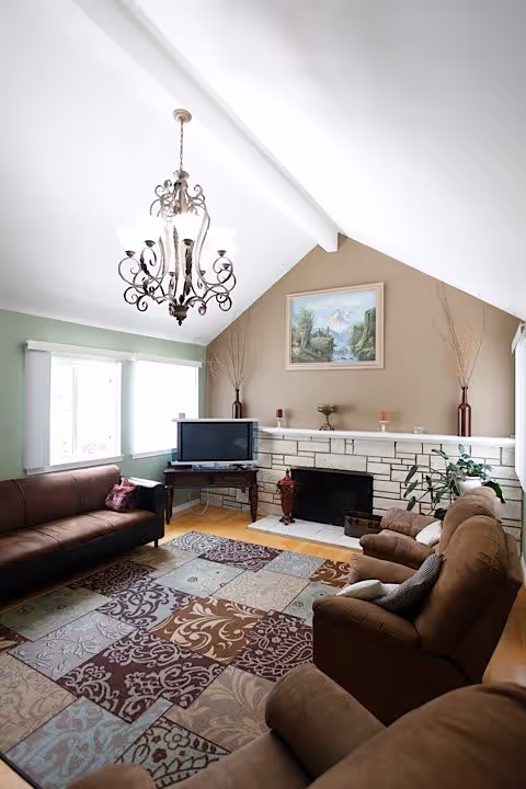 A cozy living room with a high vaulted ceiling and an ornate chandelier. The room features a brown leather sofa, two brown upholstered armchairs, a patterned area rug, a TV on a wooden stand, and a stone fireplace with decorative items on the mantel. A landscape painting hangs above the fireplace, and there are tall vases with branches on either side.