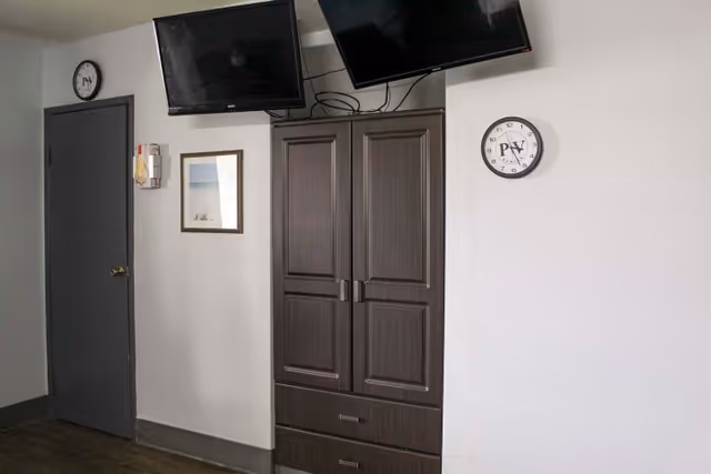 Interior wall with a dark wardrobe and drawers, two wall-mounted TVs above, two clocks marked 'PV', a framed picture, and a closed gray door.
