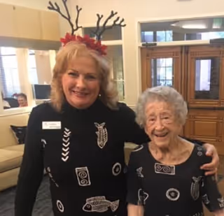 Two women smiling indoors, one wearing a black sweater with fish designs and reindeer antlers headband, the other wearing a matching black dress with fish designs. They are standing in a room with wooden doors and a beige couch in the background.