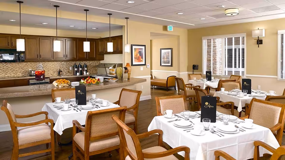 A dining room with several tables covered in white tablecloths, set with plates, cups, glasses, and silverware. Wooden chairs with cushioned seats surround the tables. In the background, there is a kitchen area with wooden cabinets, a countertop with food and drink items, and pendant lights hanging from the ceiling. The room has beige walls, windows with white blinds, and framed artwork on the walls.