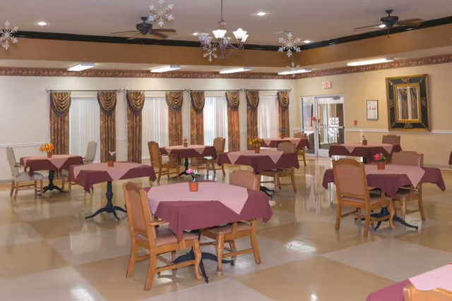 Dining room with multiple round tables draped in burgundy tablecloths, wooden chairs, small floral centerpieces, chandeliers, and draped windows.
