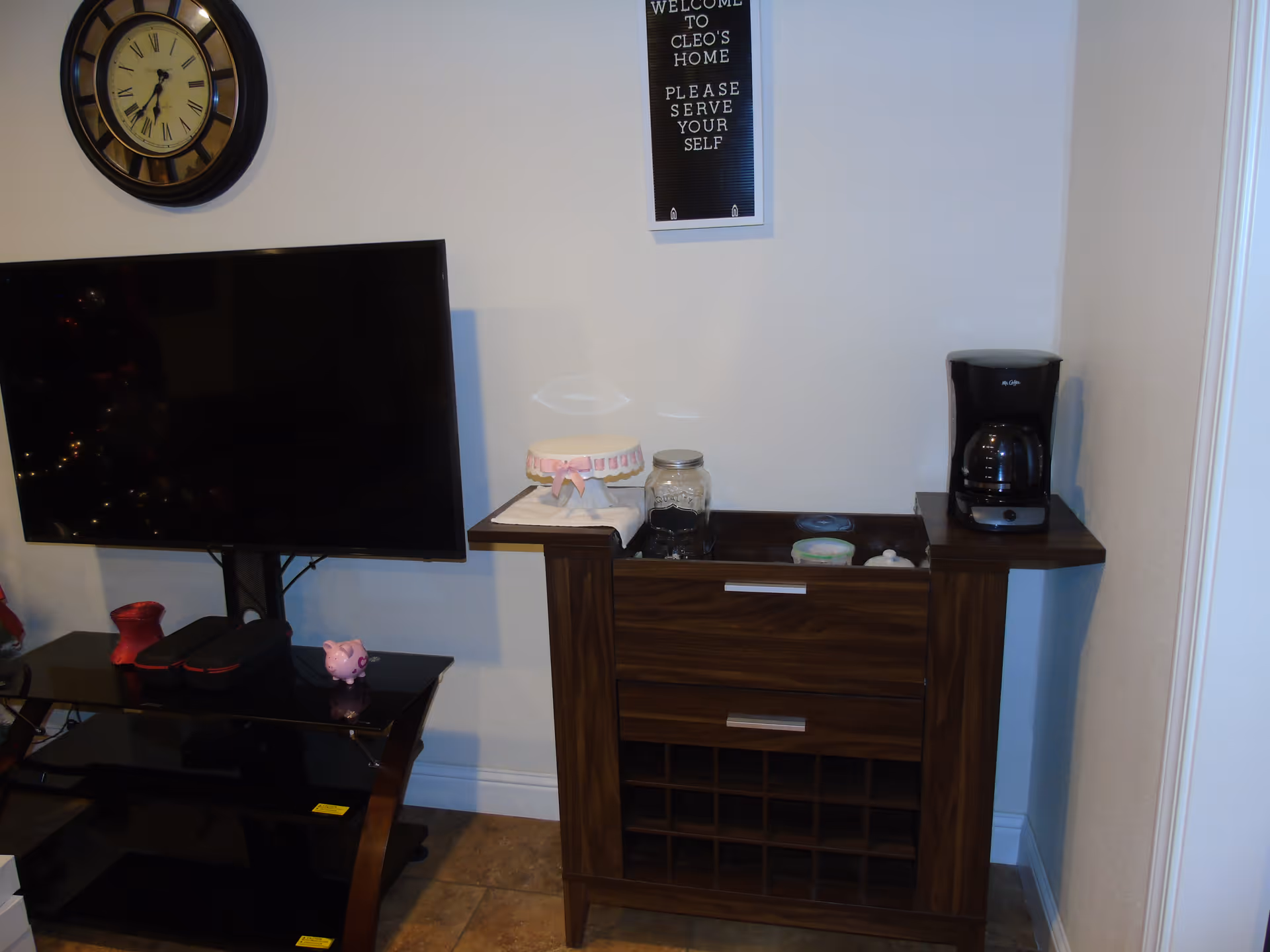 A corner of a room featuring a wall-mounted clock, a flat-screen TV on a black stand with a small pink pig figurine, and a wooden cabinet with a coffee maker on top. Above the cabinet is a sign that reads 'Welcome to Cleo's Home Please Serve Yourself.'