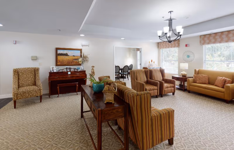 Bright senior living common room with striped armchairs, a sofa, a piano, and a wooden console table.