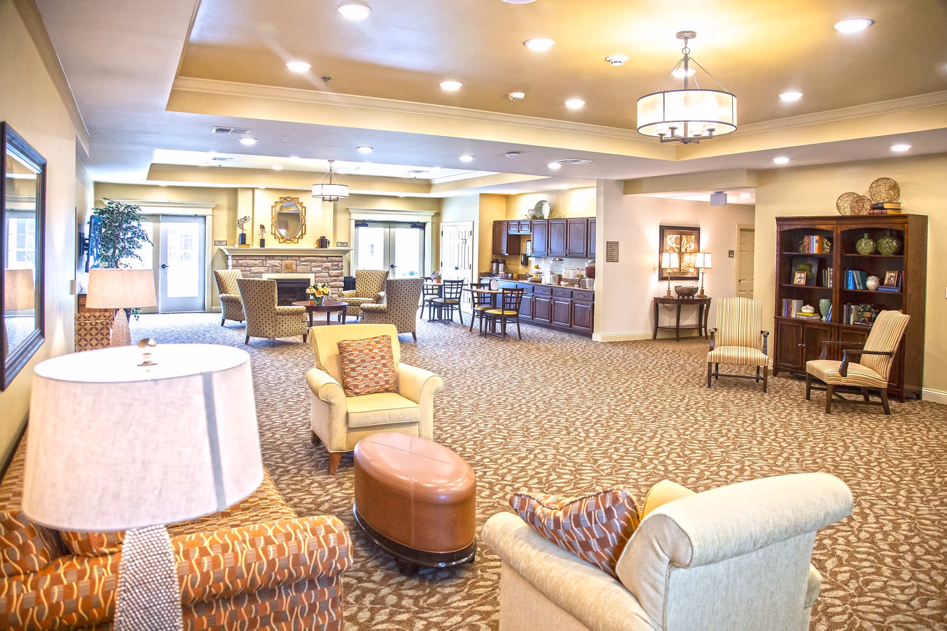 A spacious and well-lit common area in a senior living facility featuring comfortable armchairs, a sofa, a round ottoman, a fireplace with a mirror above it, a bookshelf with decorative items, and a kitchenette with tables and chairs in the background.
