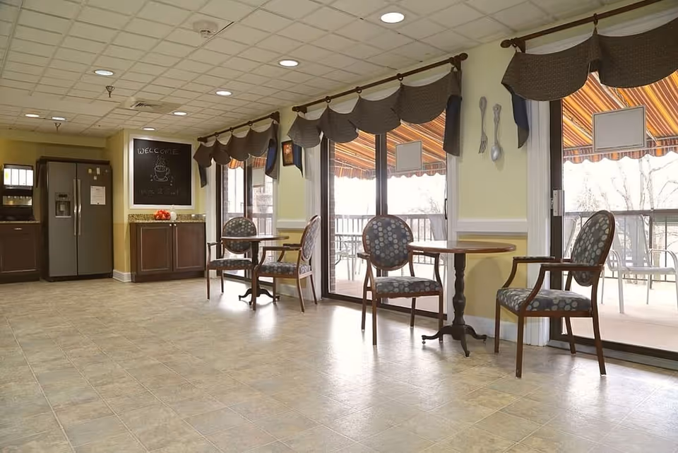 A bright dining area with round tables and cushioned chairs with patterned upholstery. Large windows with valances and striped awnings outside let in natural light. In the background, there is a kitchen area with a refrigerator, cabinets, and a chalkboard sign that says 'WELCOME' with a drawing of a coffee cup.