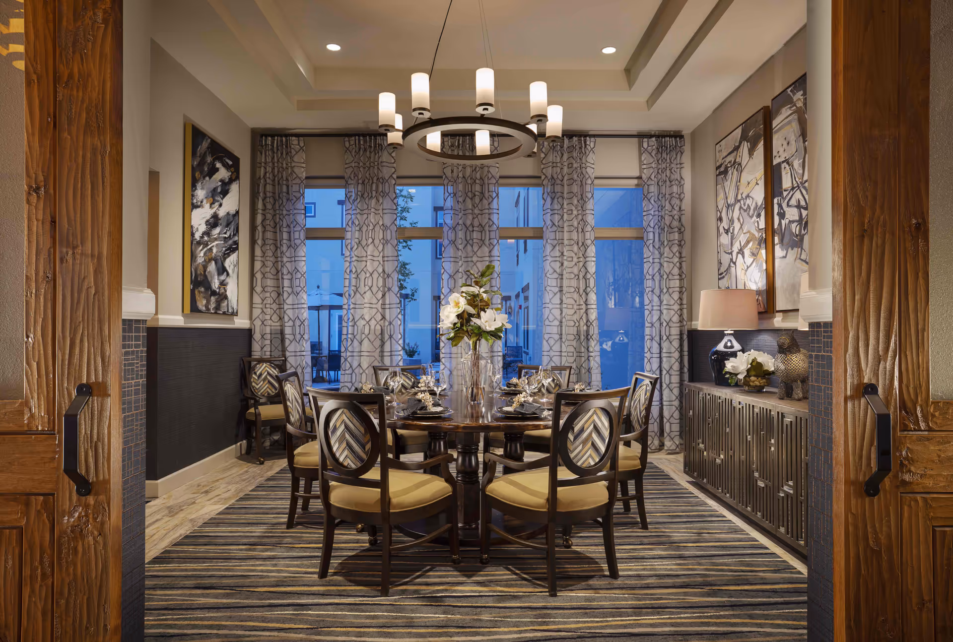 A well-lit dining room with a round wooden table set for six, surrounded by cushioned chairs with patterned backs. The room features large windows with patterned curtains, abstract artwork on the walls, a modern chandelier, and a sideboard with decorative items and a lamp. The floor has a striped carpet, and the entrance has wooden double doors with black handles.