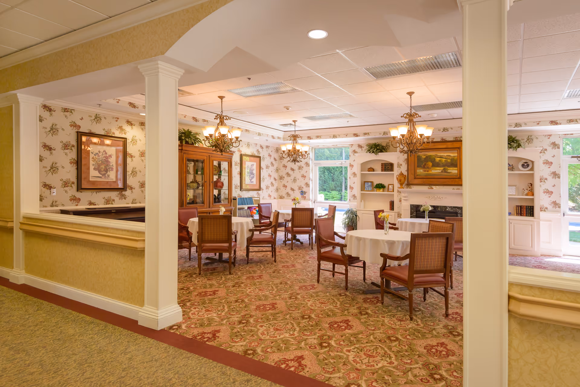 A cozy dining room in a senior living facility with round tables covered with white tablecloths and surrounded by wooden chairs. The room features floral wallpaper, a patterned carpet, chandeliers, framed artwork, a wooden cabinet, and built-in shelves with decorative items. Large windows allow natural light to brighten the space.