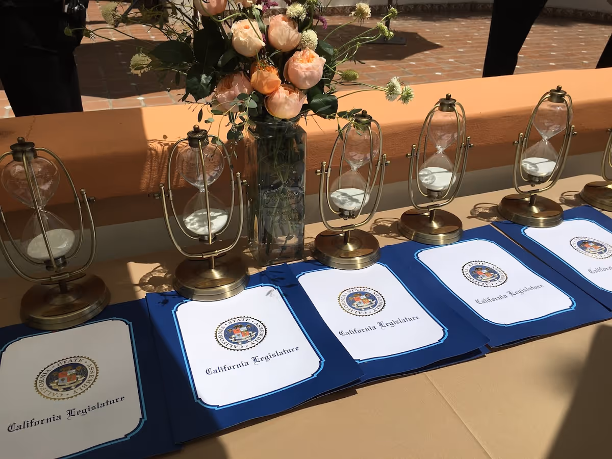 A table outdoors displaying a row of brass hourglasses, California Legislature certificates, and a vase of pink roses.
