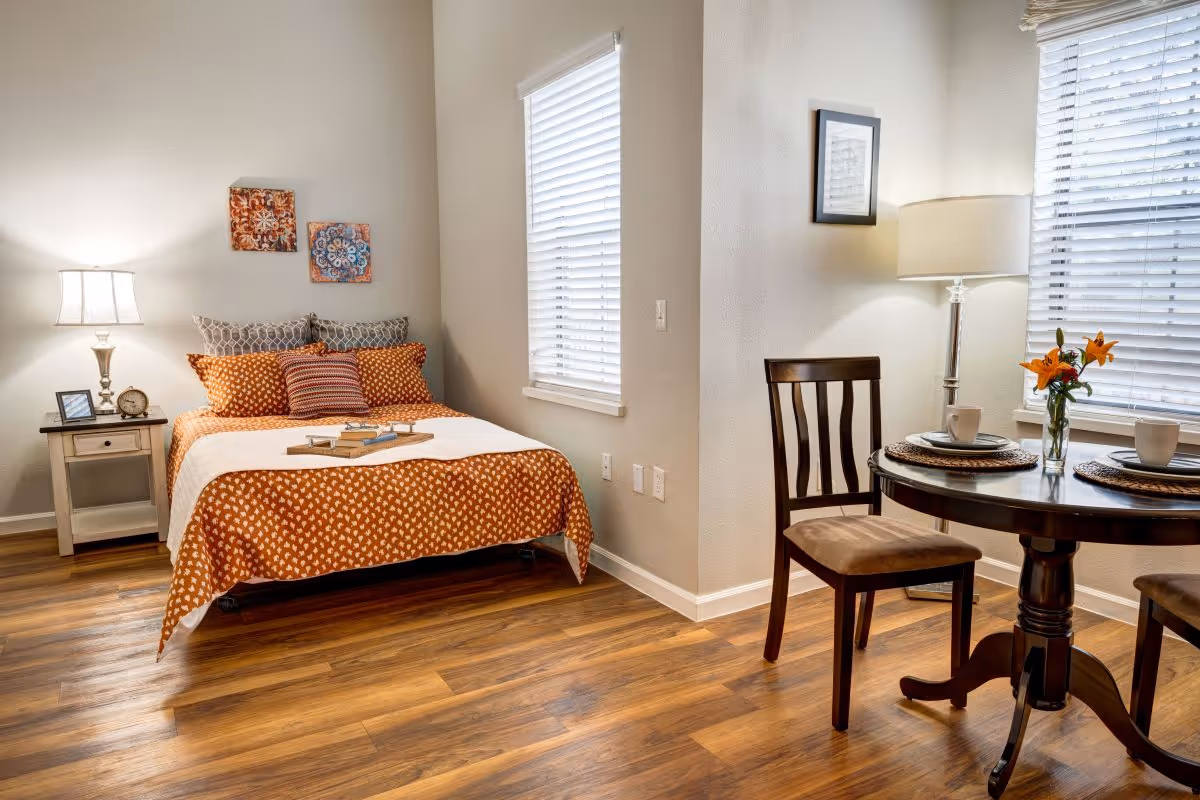 A cozy room with a bed on the left side covered with an orange and white patterned bedspread and multiple pillows. A small wooden nightstand with a lamp, clock, and picture frame is next to the bed. On the right side, there is a round wooden table set for two with placemats, cups, and a vase with orange flowers. Two wooden chairs are placed around the table. The room has wooden flooring and two windows with white blinds letting in natural light.