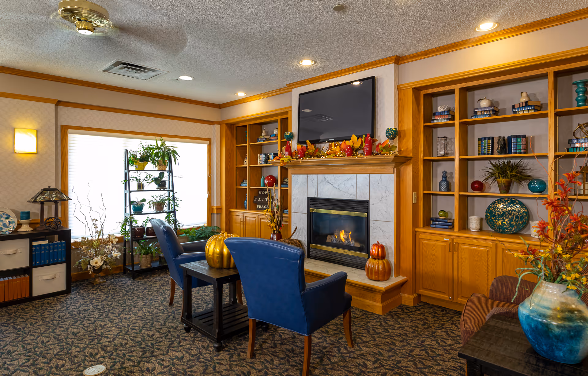 A cozy living room area in an assisted living facility featuring a lit fireplace with a TV mounted above it. The fireplace mantel is decorated with autumn-themed ornaments. Two blue upholstered chairs face the fireplace with a small wooden table between them. Built-in wooden shelves on either side of the fireplace hold various decorative items and books. A large window with a plant stand holding multiple potted plants allows natural light into the room. The carpet has a patterned design, and the walls are light-colored with wooden trim.