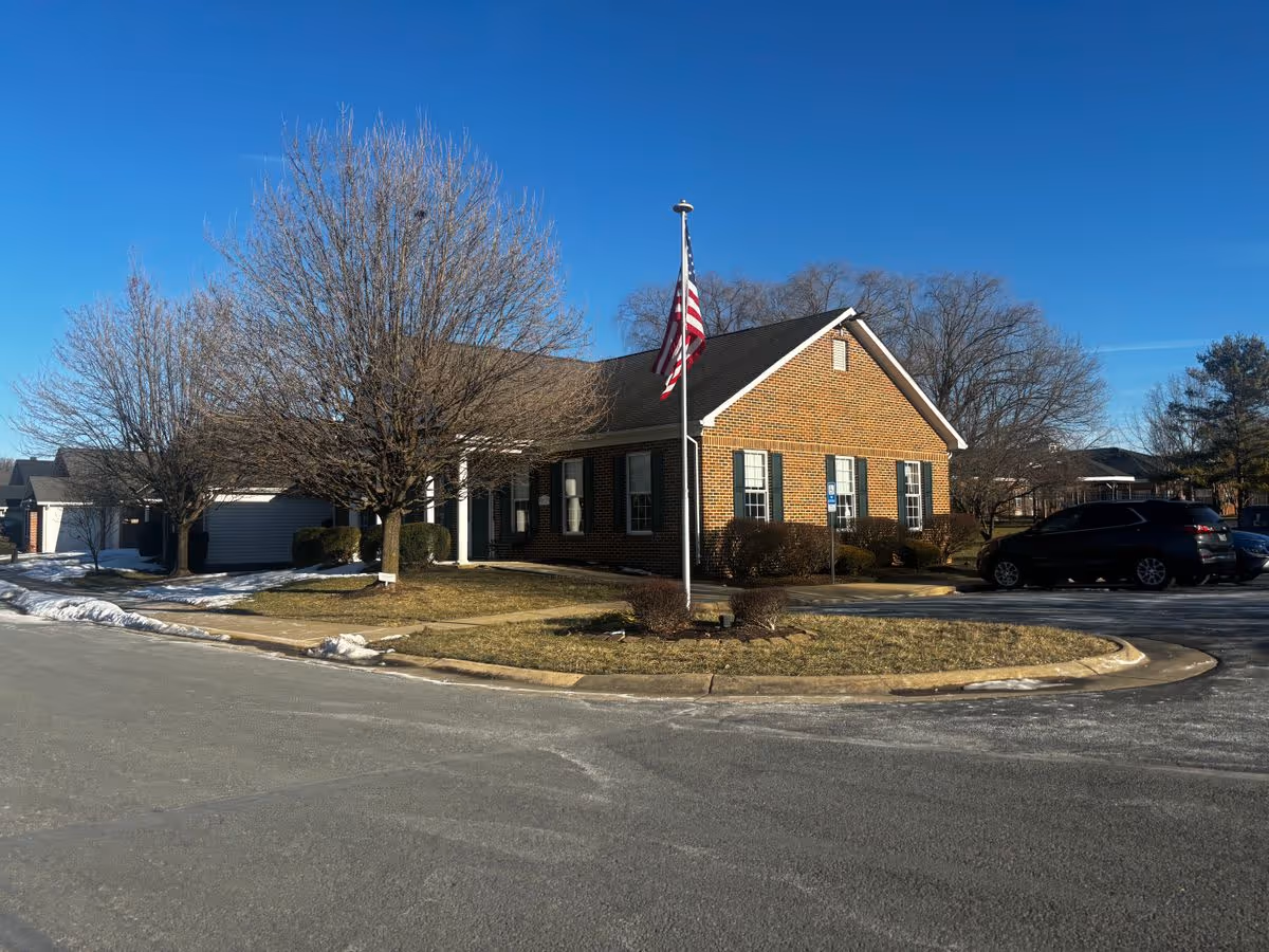 Single-story brick building with an American flag on a flagpole, leafless trees, and parked cars under a clear blue sky.