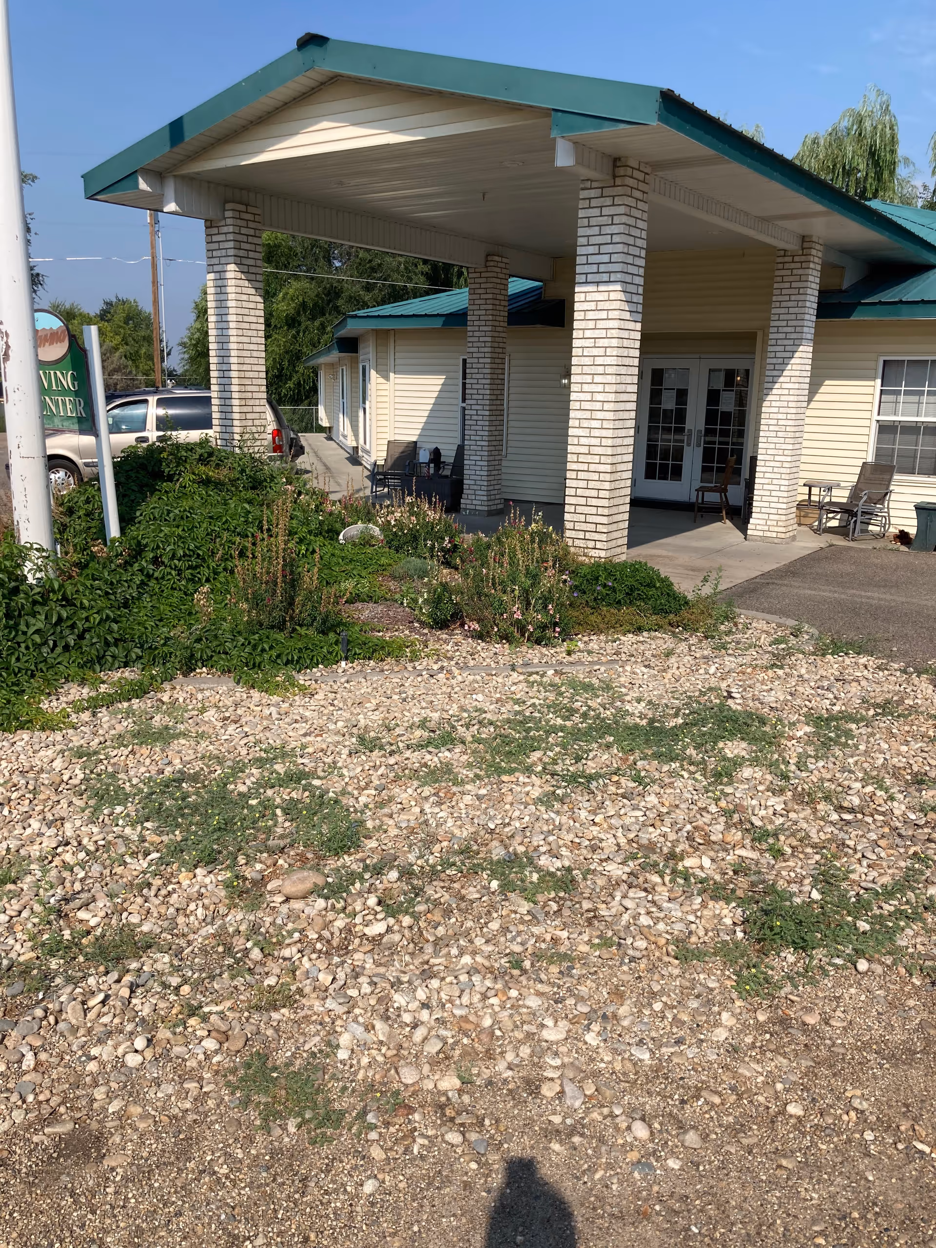 Exterior view of Parma Living Center showing the entrance with a covered porch supported by white brick columns. There are chairs on the porch and a gravel area with some greenery and plants in front. A sign partially visible on the left side reads 'Parma Living Center'.