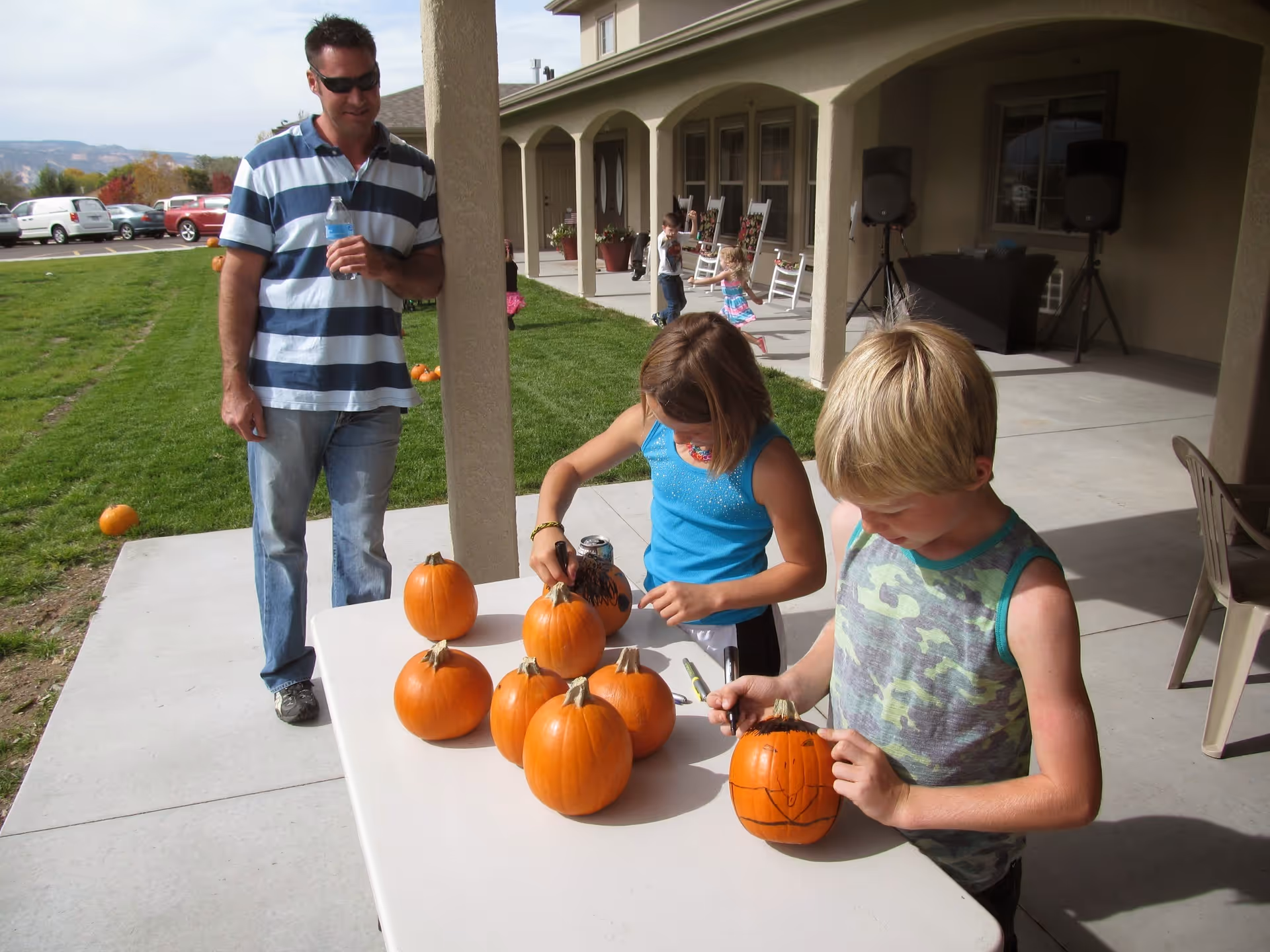 Two children are decorating small pumpkins on a white table outside a building with a covered porch. A man in a striped shirt and sunglasses is standing nearby holding a water bottle. Other children and rocking chairs are visible on the porch in the background.