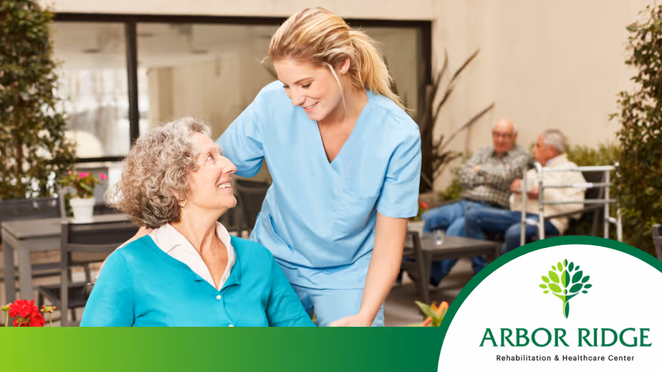 A female healthcare worker in blue scrubs smiling and interacting with an elderly woman in a turquoise sweater seated in a wheelchair in an outdoor patio area. In the background, two elderly men are sitting and talking on benches surrounded by plants.
