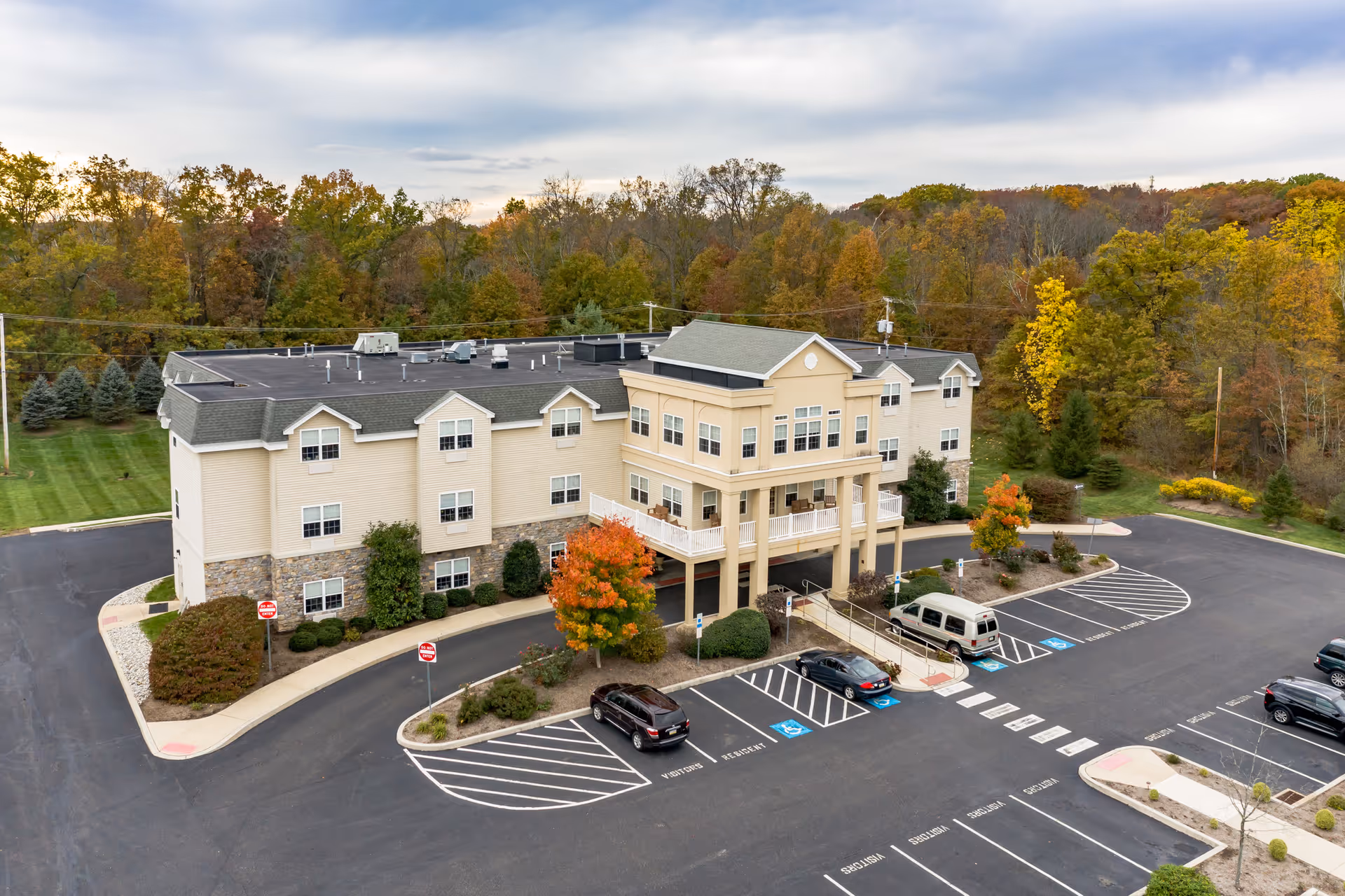 Aerial view of a three-story senior living facility building named Hidden Meadows On the Ridge surrounded by a parking lot with several cars and landscaped greenery. The building is beige with stone accents on the lower level and has a covered entrance supported by columns. Trees with autumn foliage are visible in the background.