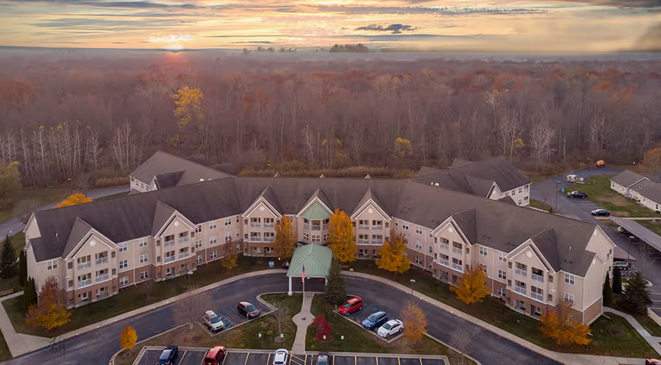 Aerial view of a large senior living facility building with multiple peaked roofs and a green-roofed entrance canopy, surrounded by a parking lot with several cars and autumn-colored trees, with a forest and a sunset in the background.