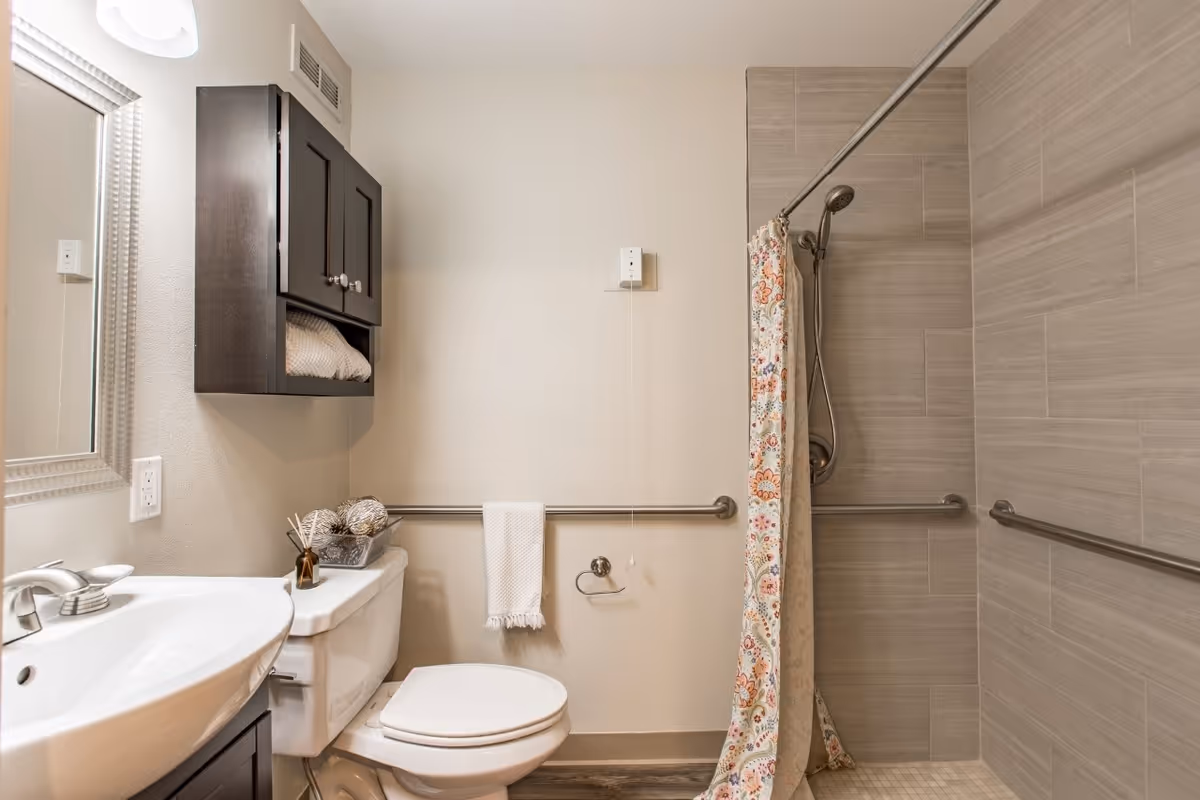 A clean and modern bathroom featuring a white sink with silver faucet, a toilet with a towel rack above it holding a white towel, a dark wooden cabinet mounted on the wall with towels inside, and a shower area with beige tiles and a floral-patterned shower curtain. The bathroom has grab bars installed for accessibility.