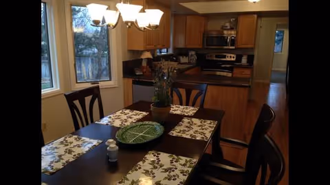 Dining area with a dark wooden table set for four with floral placemats and a green decorative plate in the center. A potted plant is also on the table. The room has large windows letting in natural light, and in the background, there is a kitchen with wooden cabinets, a microwave, stove, and countertops.
