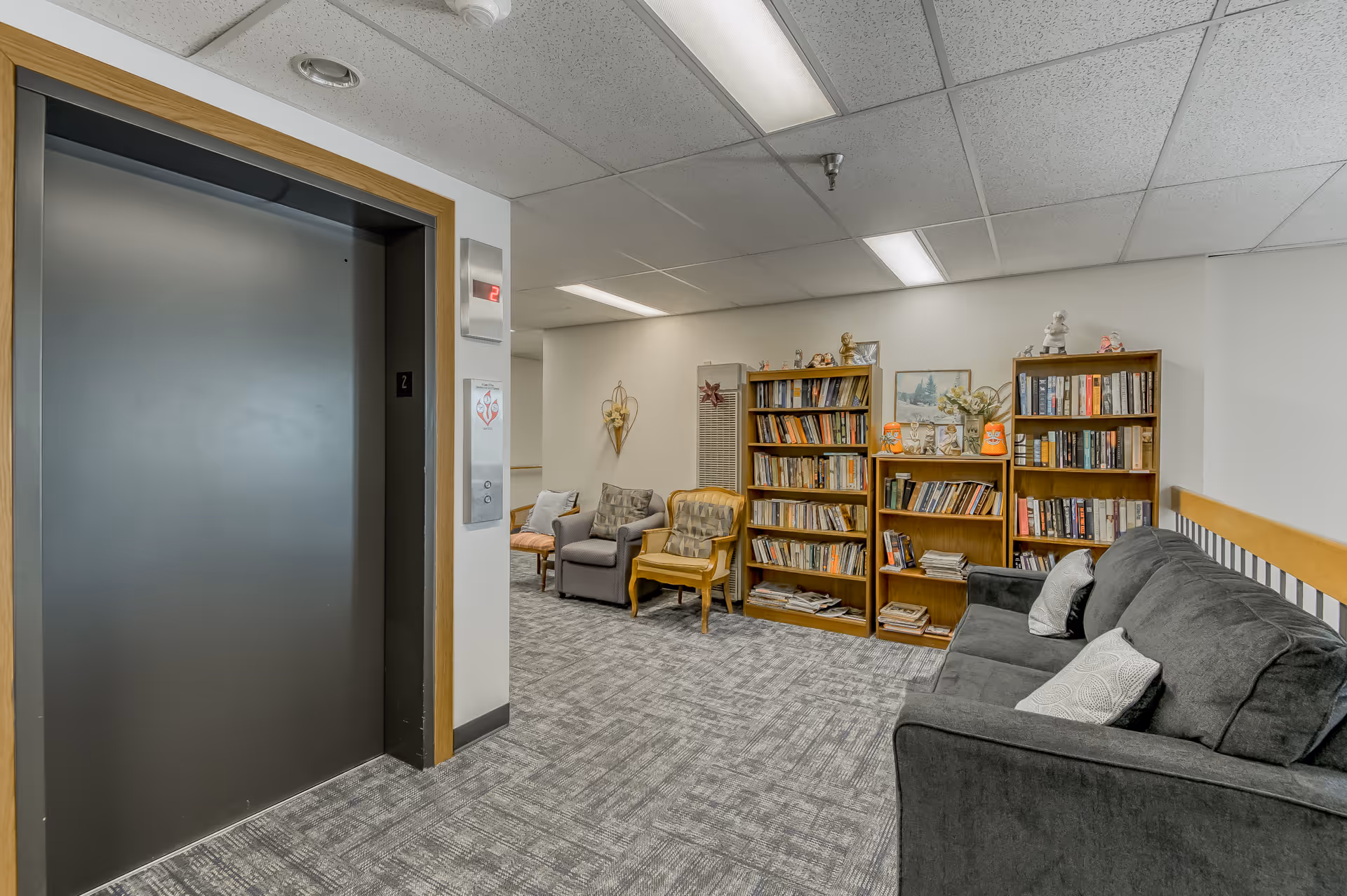 Interior view of a senior living facility hallway with a closed elevator door on the left, a seating area with a gray couch and two armchairs, and two wooden bookshelves filled with books and decorative items against the wall.