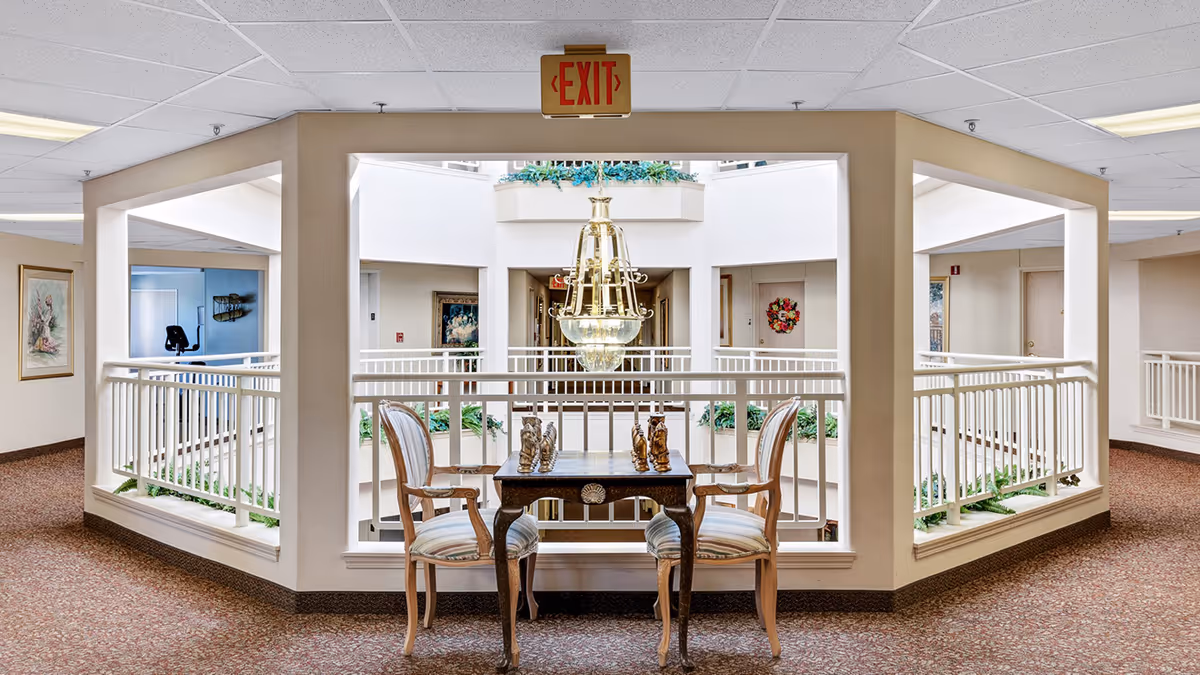 Interior view of a senior living facility hallway with a small table and two chairs set up for playing chess. The area overlooks an open atrium with a large chandelier hanging in the center. The walls are beige, and the floor is carpeted. There are framed pictures on the walls and plants around the atrium.