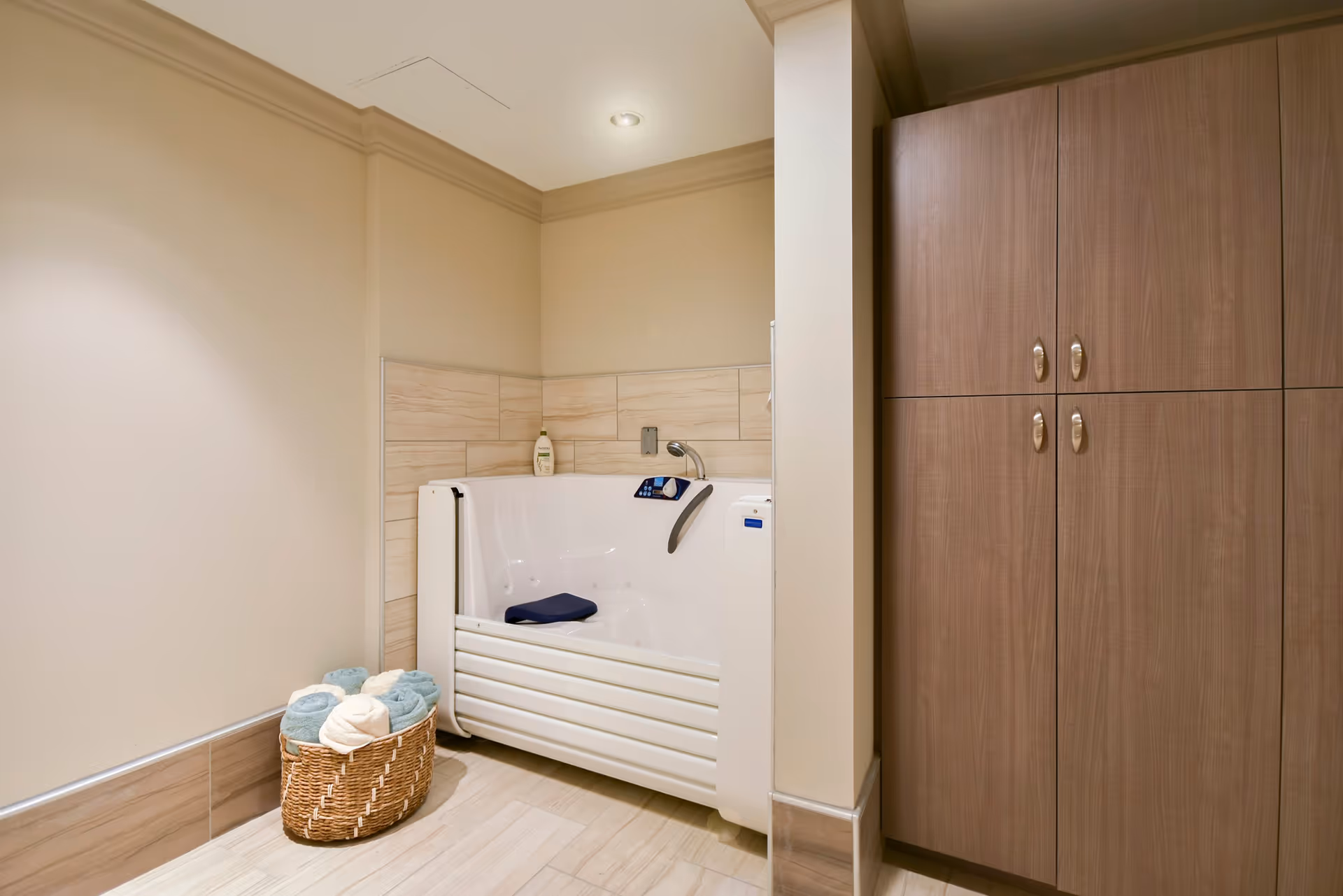 A modern bathroom corner featuring a white walk-in bathtub with a control panel and a blue cushion inside. Next to the bathtub is a wicker basket filled with rolled towels in blue and cream colors. The walls and floor are tiled in light beige tones, and there is a wooden cabinet with silver handles on the right side.