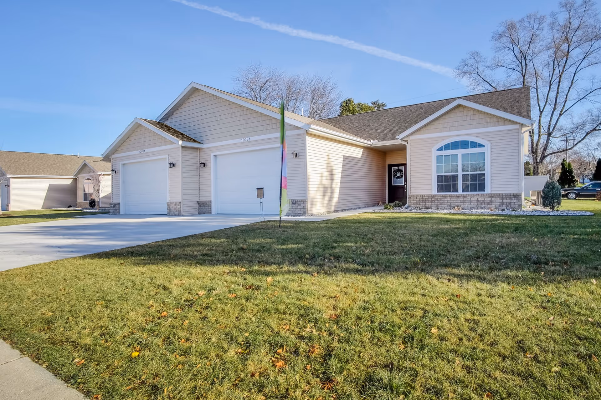 Exterior view of a single-story residential building with beige siding, two white garage doors, a large window with an arched top, and a front door with a wreath. The building is surrounded by a green lawn and a clear blue sky is visible above.