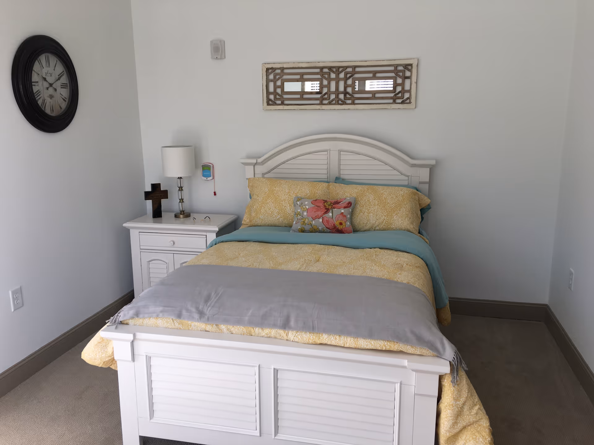 A neatly made bed with a white wooden frame and headboard, covered with yellow and teal bedding and a floral decorative pillow. To the left of the bed is a white nightstand with a lamp, a wooden cross, and a pair of glasses. A large round clock hangs on the wall above the nightstand, and a decorative rectangular wall piece is mounted above the bed. The room has white walls and beige carpet.