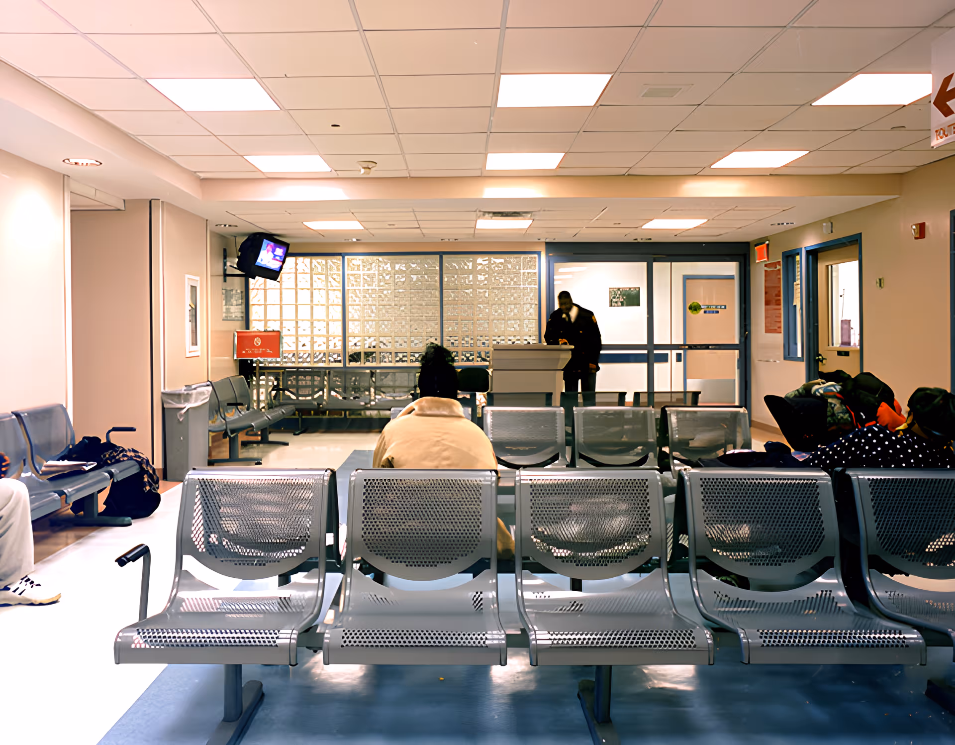 Waiting area with rows of metal chairs, a few people seated, a television mounted on the wall, and a reception desk with a person standing behind it. The room has a tiled ceiling with fluorescent lights and glass block windows in the background.
