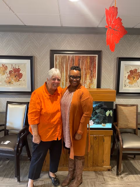 Two women standing and smiling inside a room with framed artwork on the wall behind them and an aquarium between two chairs. One woman is wearing an orange shirt and black pants, and the other is wearing a patterned dress with an orange cardigan and brown boots.