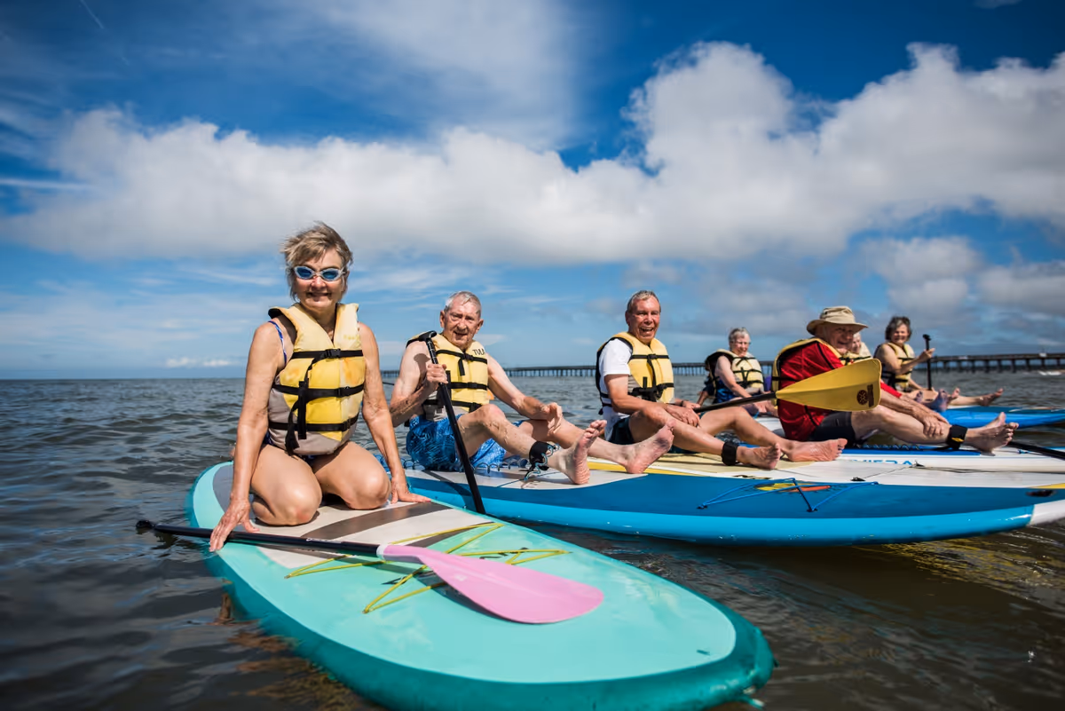A group of six elderly people wearing yellow life jackets are sitting on paddleboards in calm water under a partly cloudy blue sky. They appear to be enjoying a water activity together, with some holding paddles and smiling at the camera.