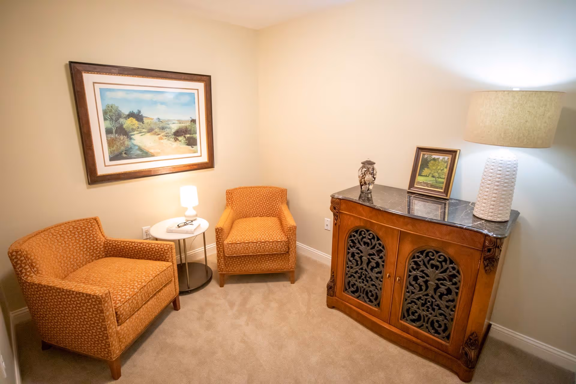 A cozy sitting area with two orange patterned armchairs, a small round white side table with a lamp and glasses on it, a wooden cabinet with ornate black metal doors, a white textured lamp, a small framed picture, and a large framed landscape painting on the wall.