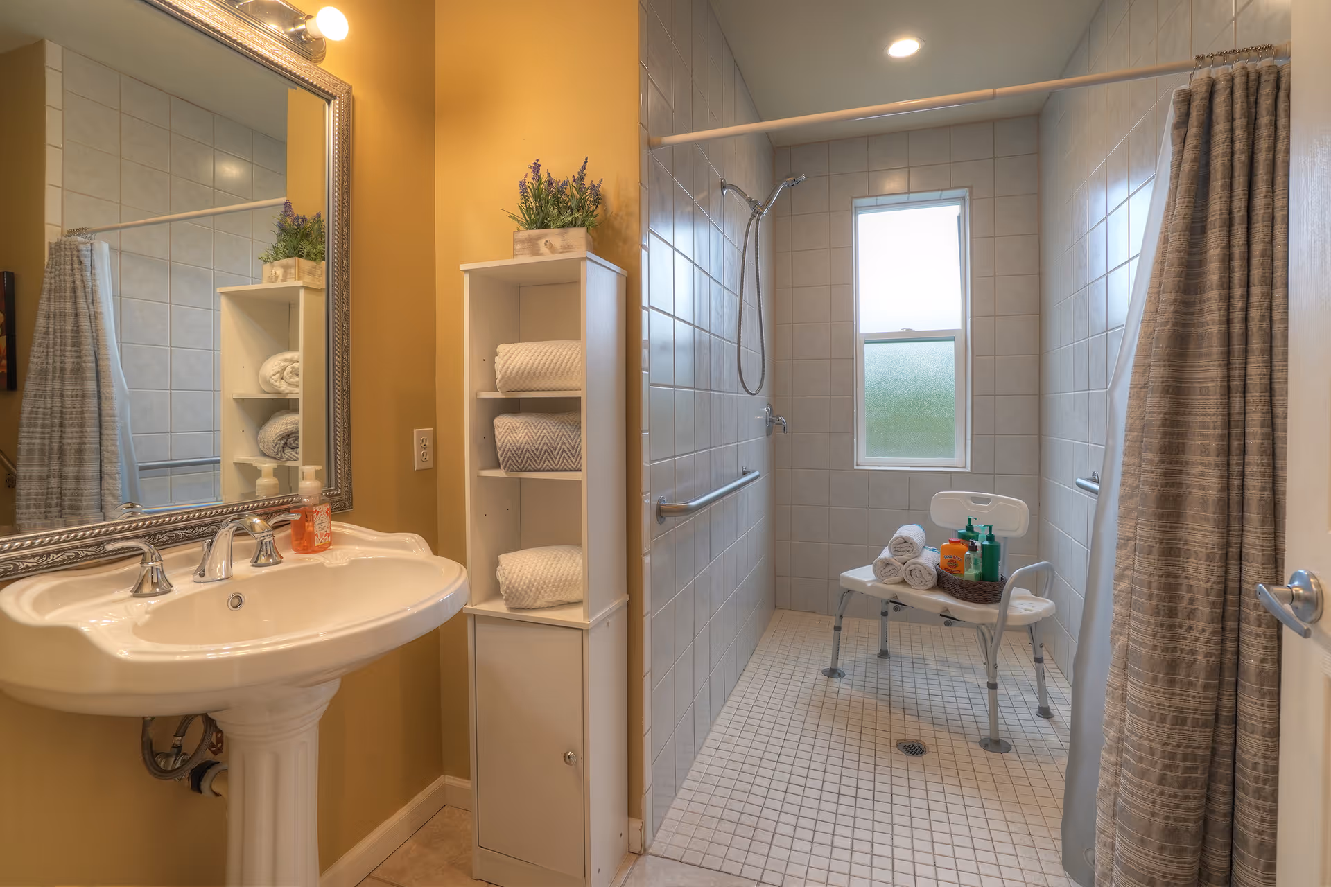 Accessible tiled bathroom with a pedestal sink, shelving with towels, and a roll-in shower with grab bars and a shower chair.