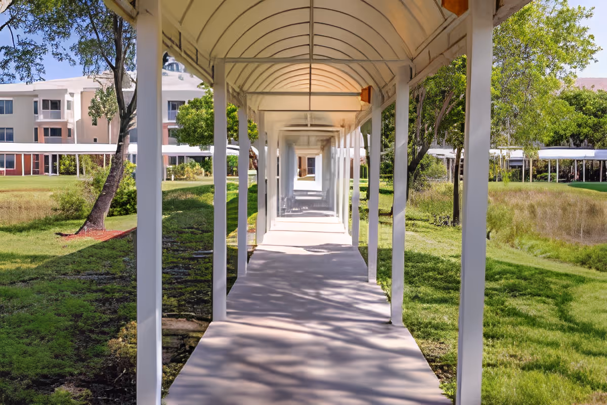 Covered outdoor walkway with white pillars and a curved roof, surrounded by green grass, trees, and a building in the background under a clear blue sky.