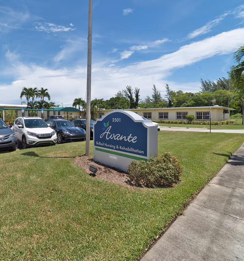 Outdoor view of Avante Skilled Nursing & Rehabilitation facility sign on a grassy area with parked cars and a building in the background under a partly cloudy blue sky.