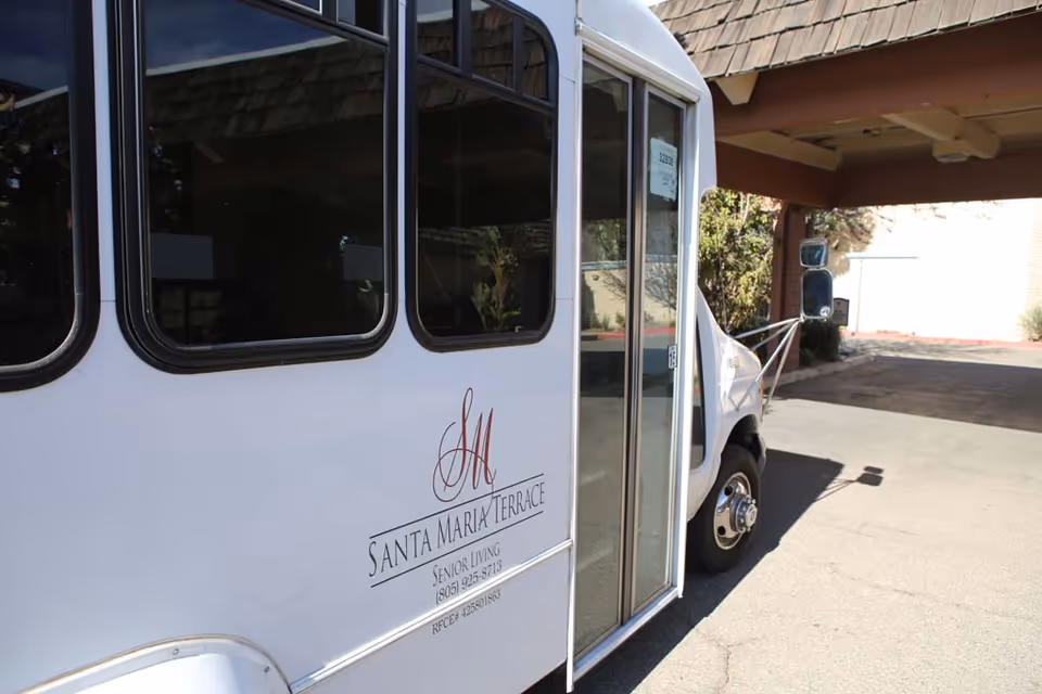 A white shuttle bus parked outside a building entrance with the logo and name 'Santa Maria Terrace Senior Living' displayed on its side.