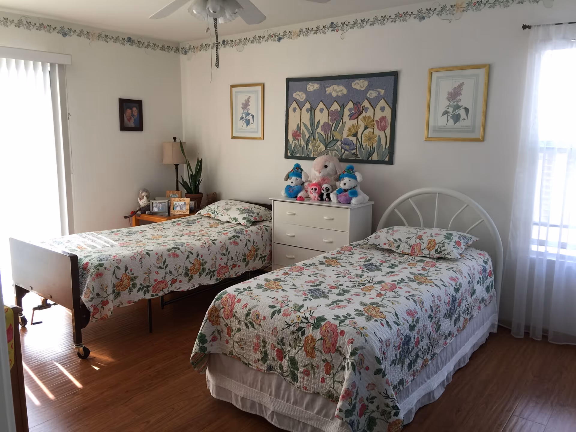 A bedroom with two single beds covered in floral bedspreads. Between the beds is a white dresser topped with stuffed animals. The room has wooden flooring, a ceiling fan, framed floral artwork on the walls, and a window with sheer white curtains letting in natural light.