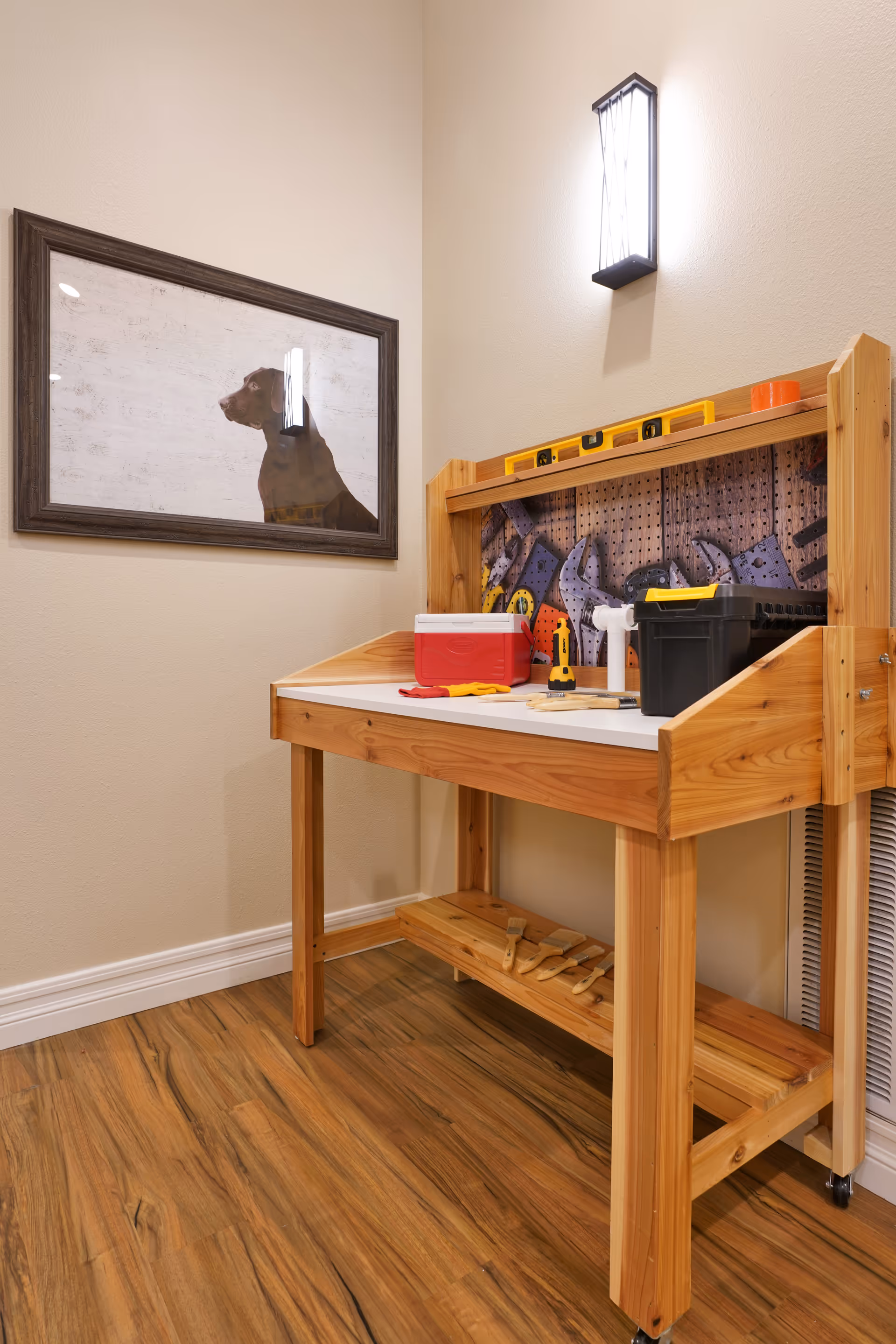 A small wooden workbench with various tools including a level, drill, and toolbox, set against a beige wall with a framed picture of a dog and a wall-mounted light fixture above.