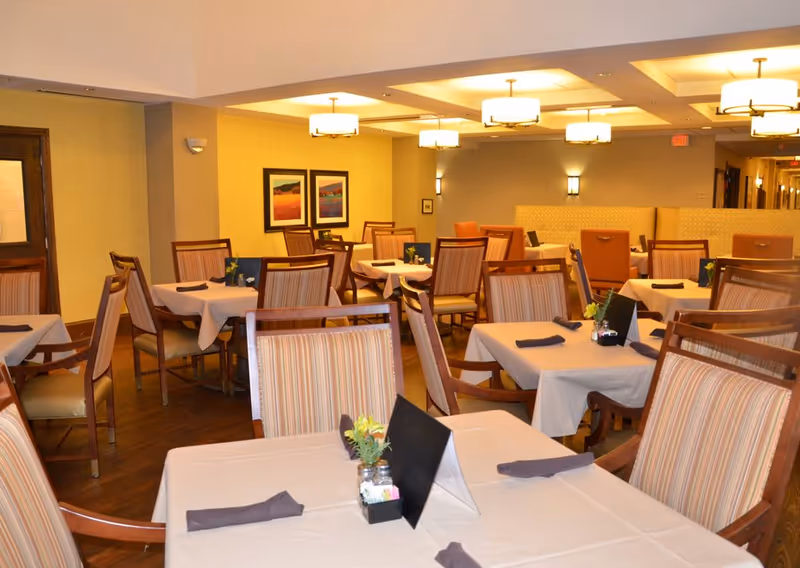 A dining room with multiple tables covered in white tablecloths, each set with folded dark napkins, small flower arrangements, and menus. The room has wooden chairs with striped upholstery, warm lighting from ceiling fixtures, and framed artwork on the walls.