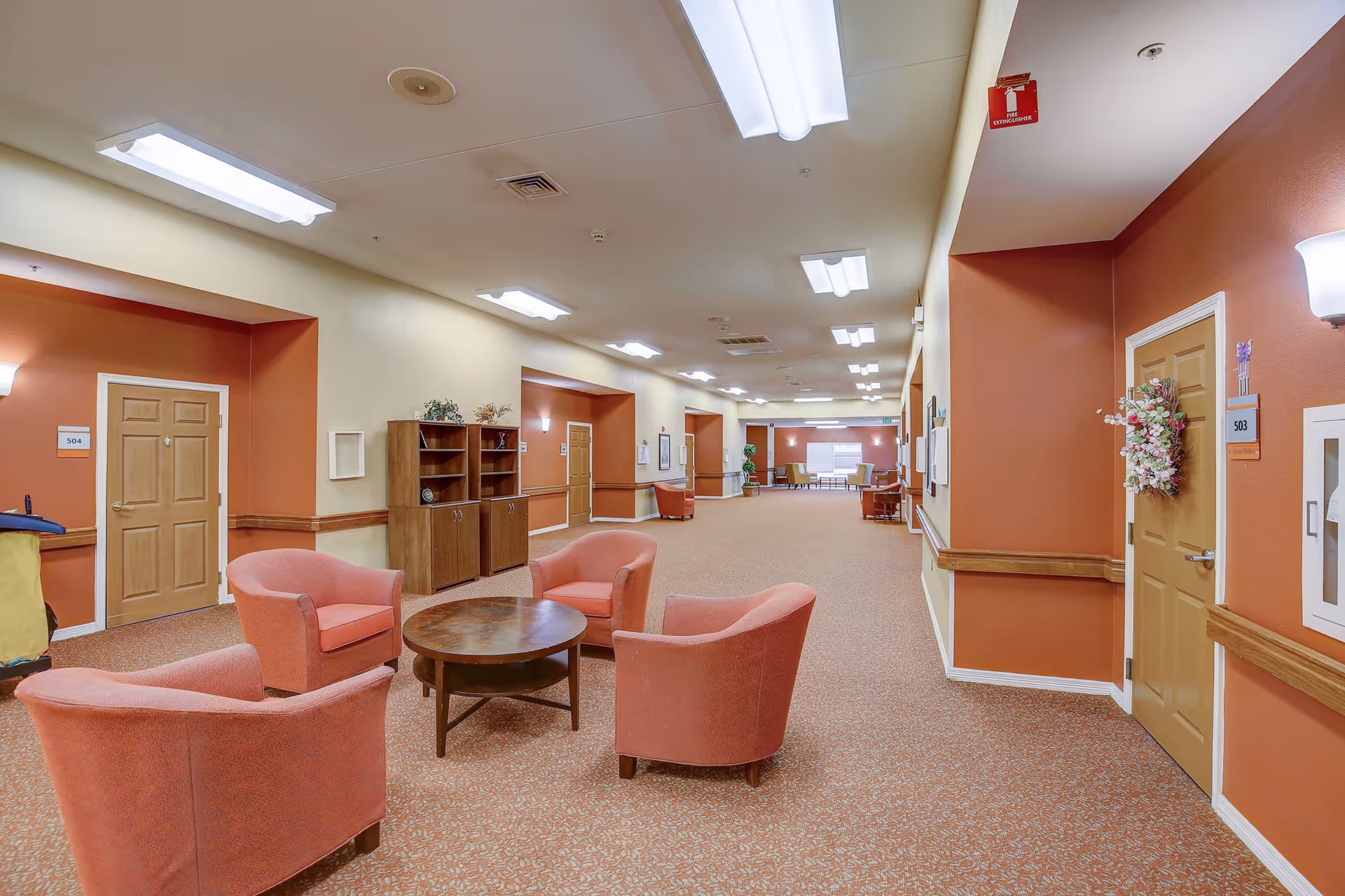 Long interior corridor of a senior living facility with a seating area of four armchairs around a coffee table and doors to resident rooms.