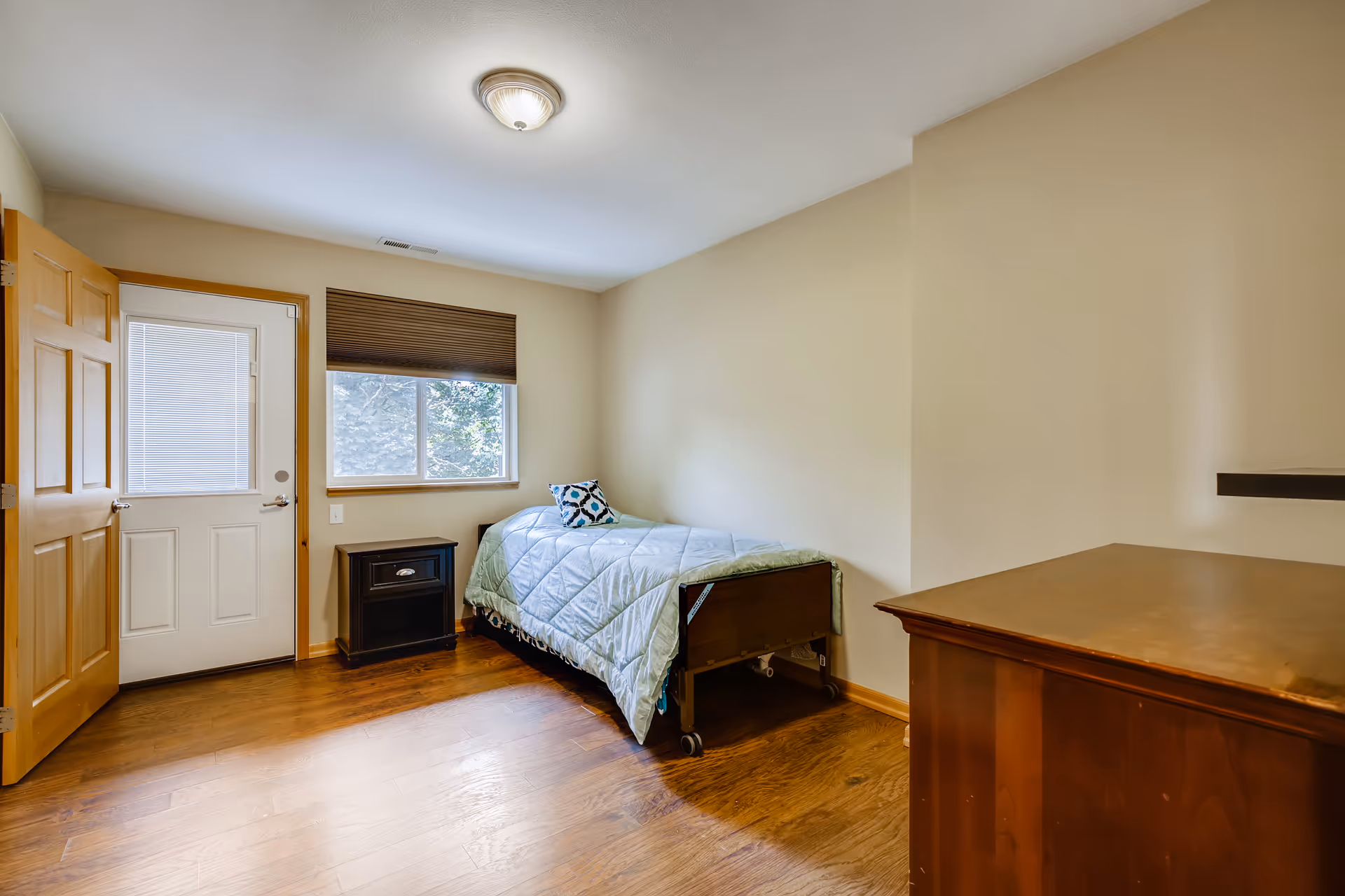 Small furnished bedroom with a single bed on casters, a nightstand, window, door, and a wooden dresser.