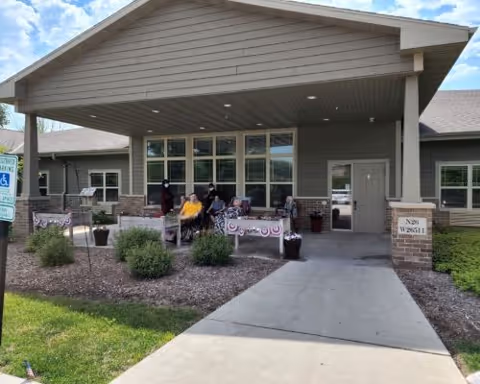 Exterior view of the entrance to Dimensions Living Pewaukee West facility with a covered porch area. Several people are seated on chairs under the porch, surrounded by small bushes and flower pots. The building has large windows and a beige siding exterior. A sidewalk leads up to the entrance, and a handicap parking sign is visible on the left side.