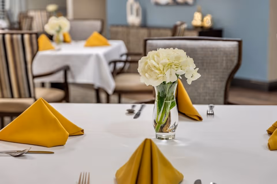 A dining table set with a white tablecloth, yellow folded napkins, silverware, and a clear glass vase holding white flowers. In the background, there are more tables and chairs with similar settings in a softly lit dining room.