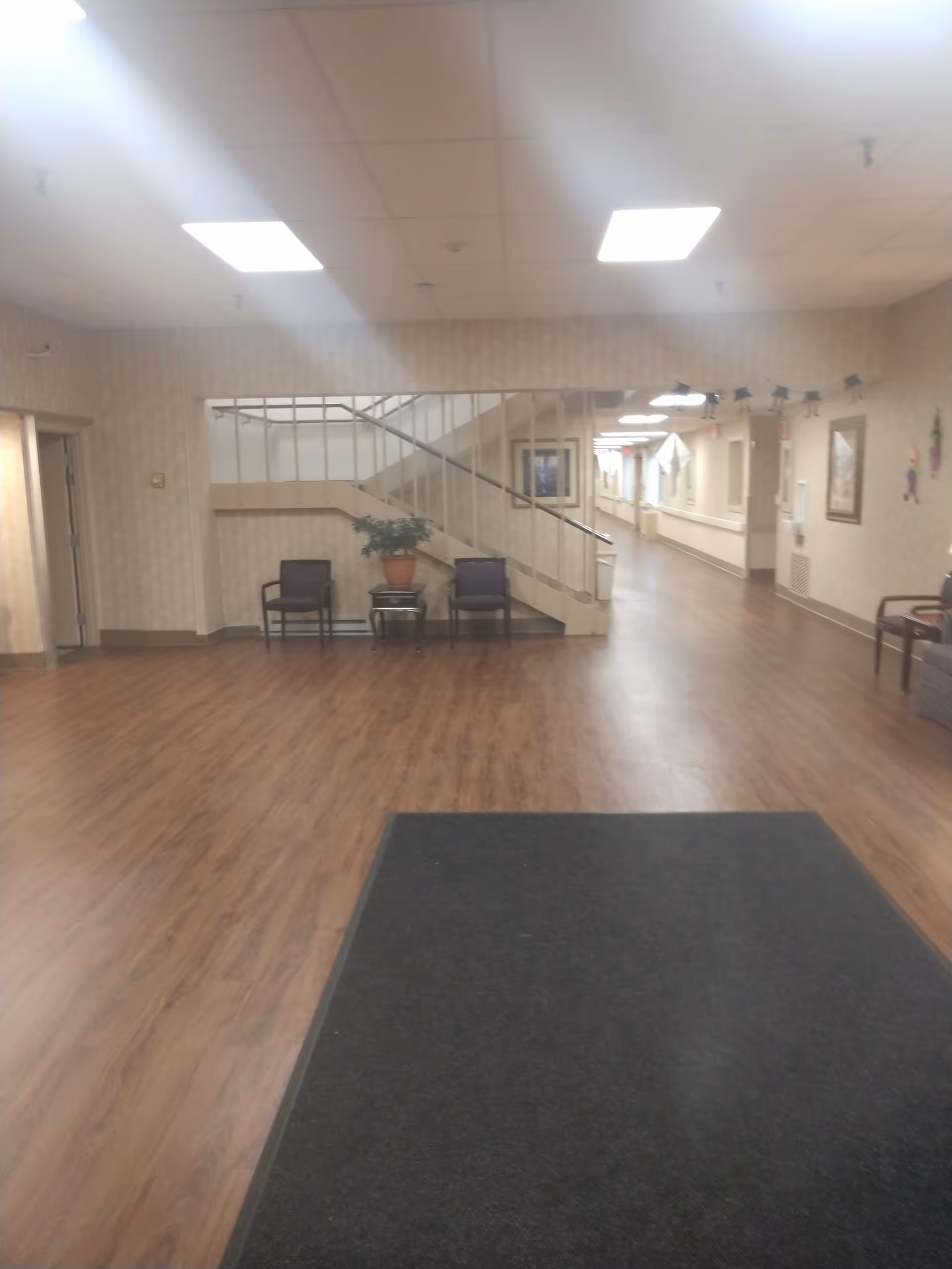 Interior view of a senior living facility hallway with wood flooring, beige walls, and ceiling lights. There is a staircase with a railing in the center background, two chairs and a small table with a potted plant underneath the stairs. The hallway extends to the right with framed pictures on the walls and additional chairs along the corridor.